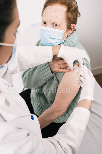 From above of crop medic in uniform with syringe vaccinating senior woman in mask during coronavirus pandemic in clinic — Stock Photo
