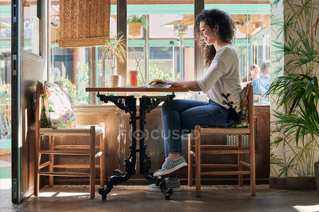 Vista lateral da funcionária étnica trabalhando no netbook à mesa com delicioso sanduíche de bagel vegetariano e bebidas — Fotografia de Stock