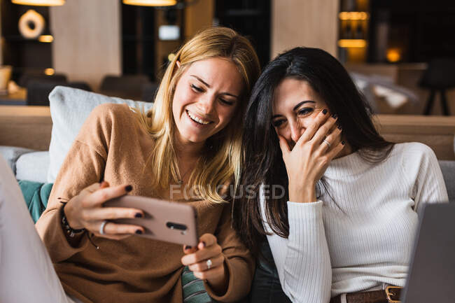 Casal de mulheres homossexuais sentadas no sofá e assistindo vídeo engraçado no telefone celular enquanto ri e se diverte — Fotografia de Stock