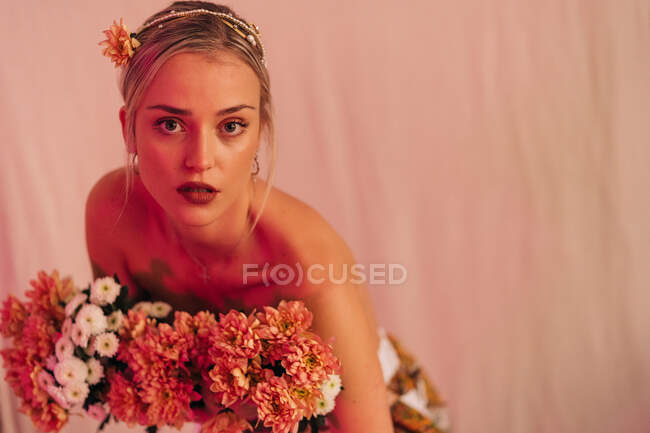 Encantadora joven romántica con hombros desnudos hembra con ramo de flores frescas en pie sobre fondo beige iluminado con luz de neón - foto de stock