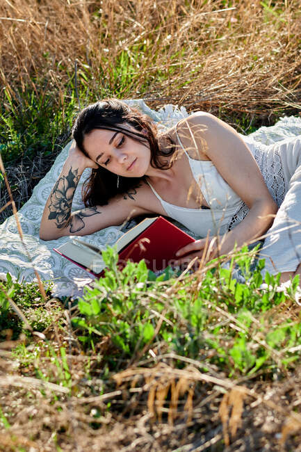 Мрійлива чарівна брюнетка в білій сукні лежить на польовому лузі і читає книгу на сонячному світлі — стокове фото