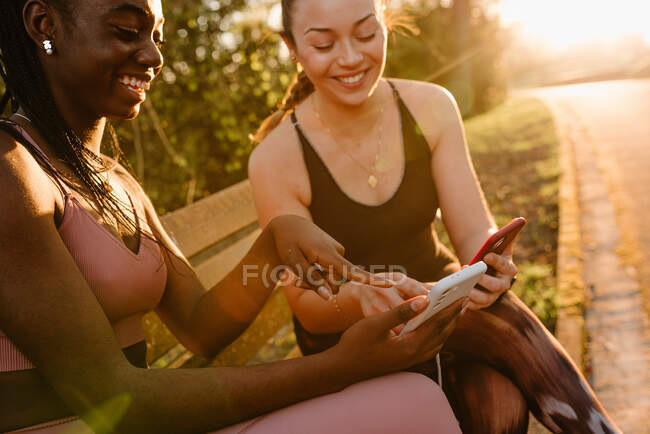 Alegres atletas multirraciales femeninas en ropa deportiva sentadas en el banco del parque y usando teléfonos móviles juntas después del entrenamiento al atardecer - foto de stock