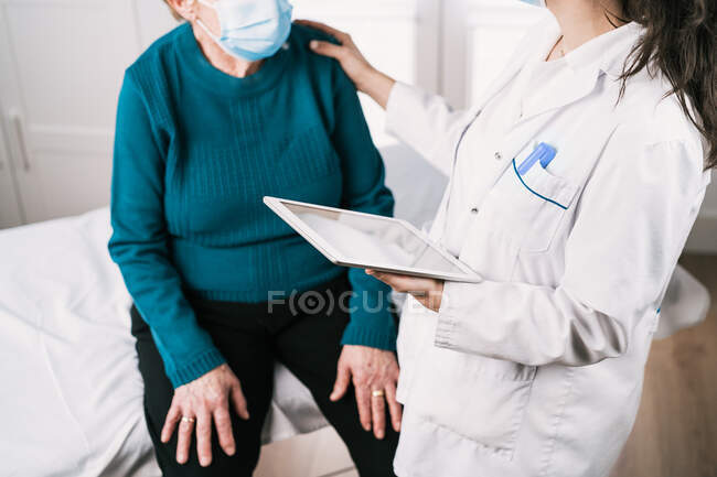 Обрезанная неузнаваемая женщина-медик в форме с планшетом, разговаривающая со старшей женщиной в стерильной маске по консультации во время пандемии ковида 19 — стоковое фото