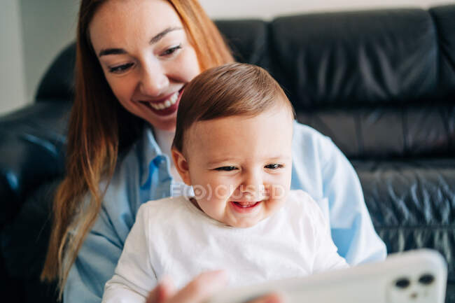 Щаслива молода мати і чарівне усміхнене дитя дивляться смішне відео на мобільному телефоні, сидячи на затишній дивані вдома. — стокове фото