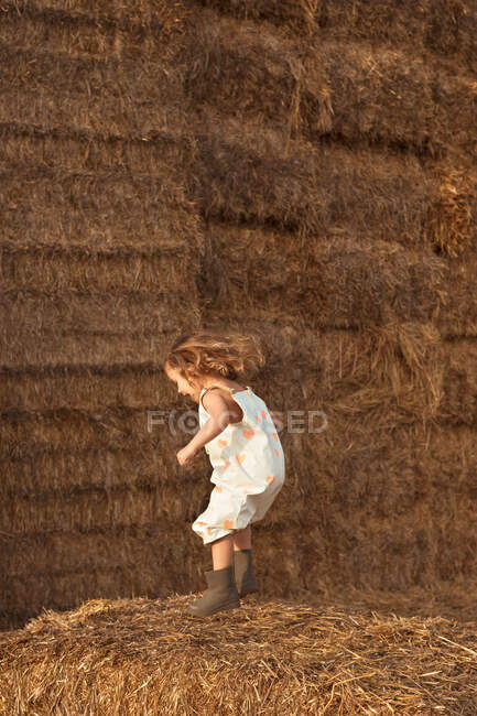Vista laterale del bambino spensierato in tuta saltando sulla balla di paglia nella giornata di sole in campagna — Foto stock