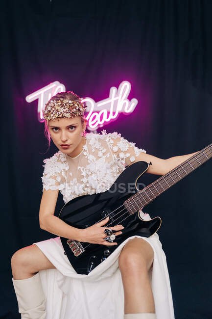 Energetic rebellious young woman in elegant white bridal dress and wreath with guitar in hand making horn gesture in studio with neon inscription — Stock Photo
