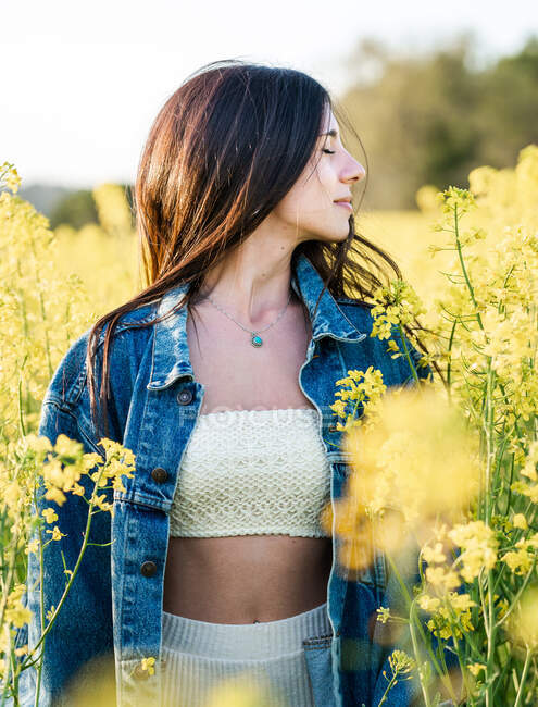 Contenuto giovane femmina in giacca di jeans in piedi con gli occhi chiusi sul campo di colza fragrante in fiore in una chiara giornata di sole — Foto stock