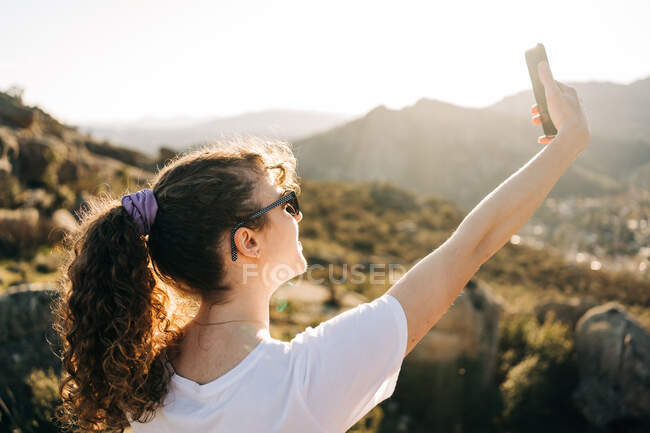 Vista posteriore di felice giovane viaggiatore donna con lunghi capelli scuri in abiti casual e occhiali da sole scattare selfie sul telefono cellulare durante il viaggio escursionistico in terreno montagnoso — Foto stock