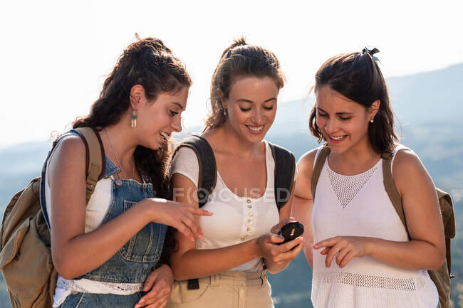 Jóvenes viajeros jóvenes felices en ropa de verano usando brújula juntos mientras están de pie en un exuberante terreno montañoso soleado - foto de stock