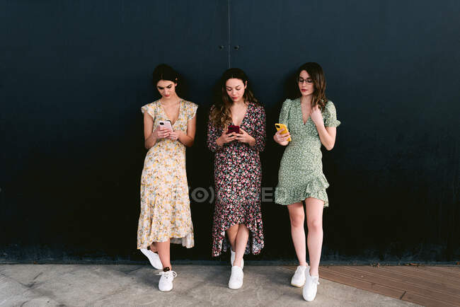 Junge beste Freundinnen in trendiger Kleidung mit Mobiltelefonen stehen auf städtischem Gehweg gegen Wand — Stockfoto