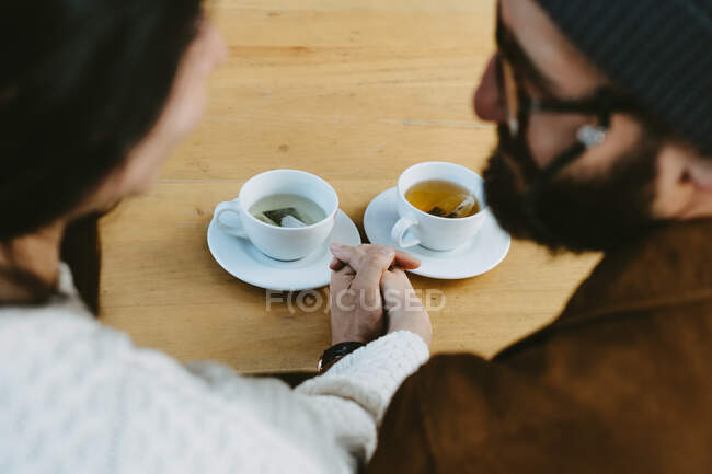 Вид зверху врожаю невпізнавана пара тримає руки, сидячи за столом з чашками гарячого чаю — стокове фото