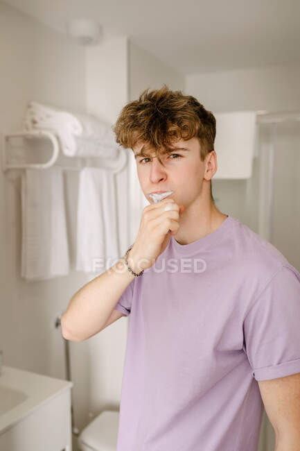 Auto asegurado joven macho millennial con el pelo de jengibre en ropa casual cepillarse los dientes y mirando a la cámara mientras está de pie en el baño en la mañana soleada - foto de stock