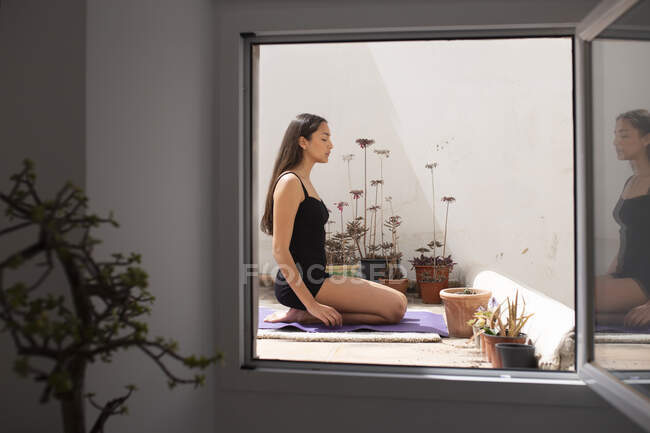 Donna a piedi nudi a tutta lunghezza seduta su Thunderbolt Posa sul tappetino yoga con gli occhi chiusi sul balcone soleggiato e meditando — Foto stock
