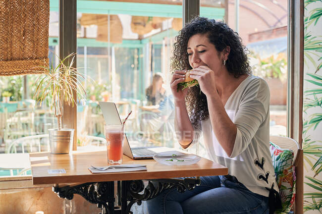 Zufriedene Fernarbeiterin mit leckerem vegetarischem Bagel-Sandwich gegen Netbook und erfrischendem Getränk im Restaurant — Stockfoto