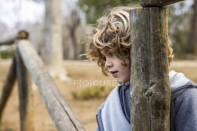 Cute child in casual outfit on old wooden fence looking away near trees in the park — Photo de stock