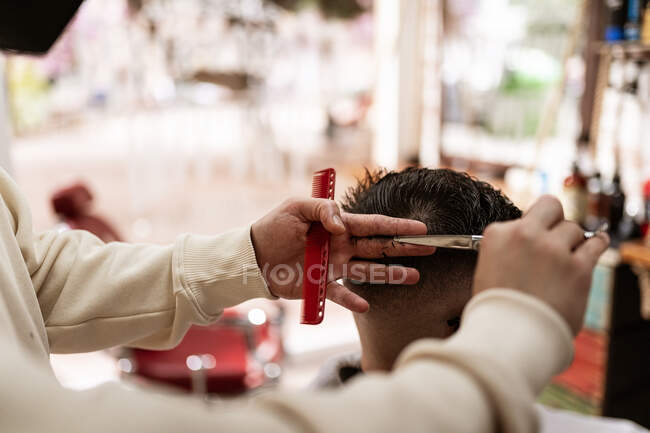 Crop anonymous male stylist with trimmer cutting hair of client in cape in barbershop — Photo de stock