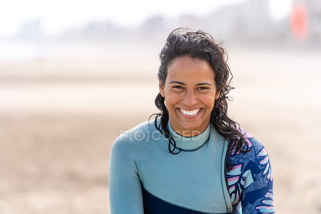 Kiter feminino étnico feliz em wetsuit com equipamento kitesurfing olhando para a câmera na praia do oceano arenoso — Fotografia de Stock
