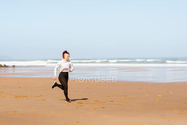 Vista lateral da jogger fêmea ativa correndo na costa oceânica durante o treinamento no verão — Fotografia de Stock