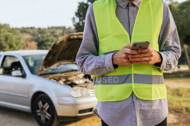 Crop unrecognizable male driver in green safety vest using mobile phone and asking for help after car accident on country road — Stock Photo