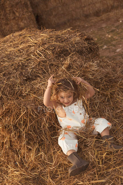 From above cheerful adorable child in overalls playing with hay lying down on straw bales in countryside — Stock Photo