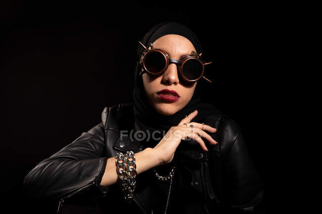 Jovem muçulmana confiante em capa de couro lenço de cabeça e óculos de sol criativos sentados contra fundo preto em estúdio — Fotografia de Stock