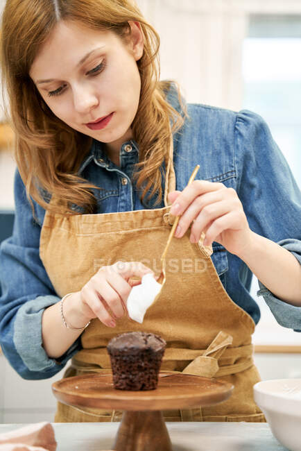 Улыбающаяся молодая женщина в фартуке со сладким кремом на венчике, смотрящая вниз во время приготовления пищи дома — стоковое фото