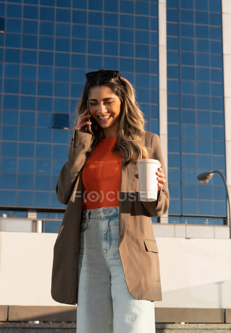 Веселая молодая женщина в стильной одежде с горячим напитком, чтобы пойти и поговорить по мобильному телефону глядя на землю в городе — стоковое фото
