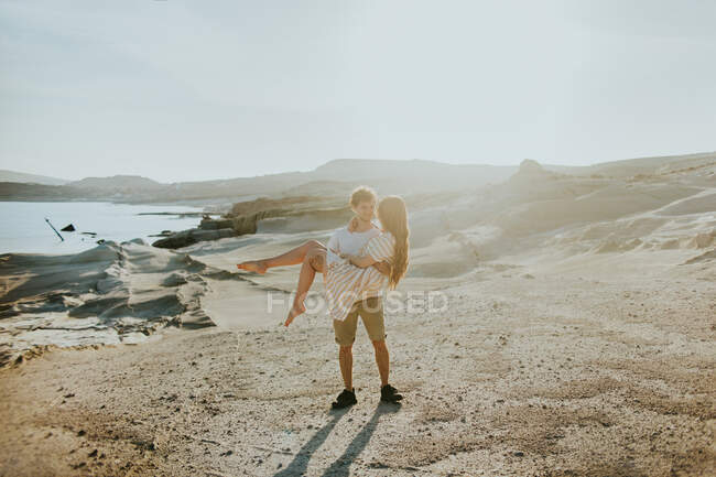 Comprimento completo jovem macho carregando namorada serena em vestido de verão elegante, enquanto em pé na costa rochosa áspera no dia ensolarado — Fotografia de Stock
