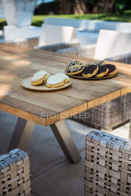 Сверху разнообразные вкусные пончики на тарелках подаются на деревянном столе возле плетеных кресел из ротанга в тропическом саду в солнечный летний день — стоковое фото