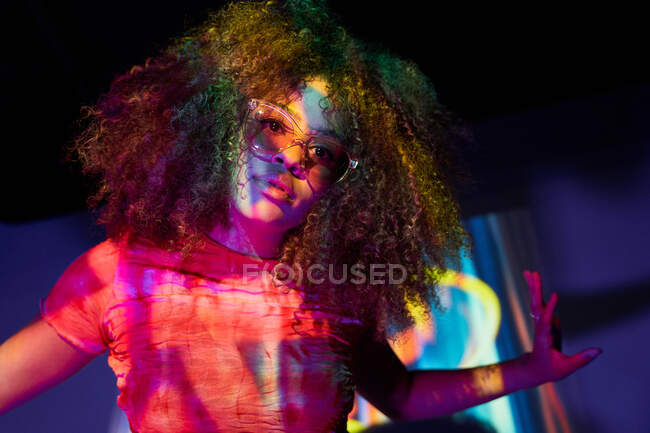 Ritratto di giovane donna afroamericana attraente in occhiali da sole alla moda guardando la fotocamera mentre in piedi in luci al neon — Foto stock