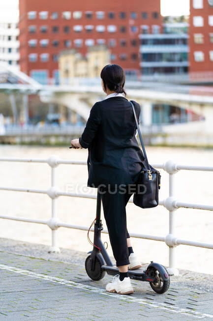 Vista posteriore di imprenditrice etnica irriconoscibile in abbigliamento formale cavalcando scooter elettrico sulla passerella in città — Foto stock
