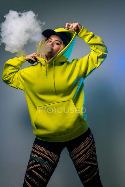 Selbstbewusste Hipsterin im Kapuzenpulli raucht E-Zigarette im Studio auf grauem Hintergrund und blickt in die Kamera — Stockfoto