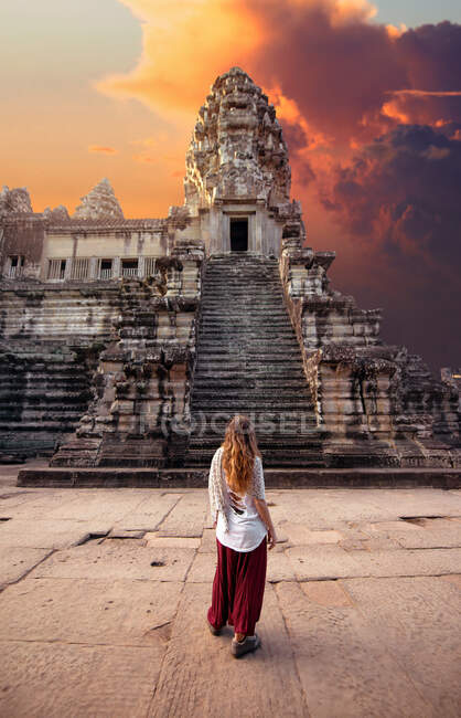 Back view of unrecognizable female tourist admiring old masonry worship exterior with staircase under cloudy sky at sundown in Cambodia — Stock Photo