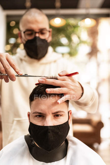 Male hairstylist in eyeglasses making haircut to adult client in hairdressing salon during COVID 19 pandemic — Foto stock