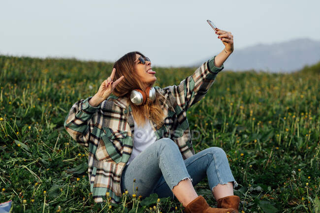 Young female in sunglasses with headphones showing peace gesture and sticking tongue out while taking self portrait on cellphone and sitting on meadow - foto de stock
