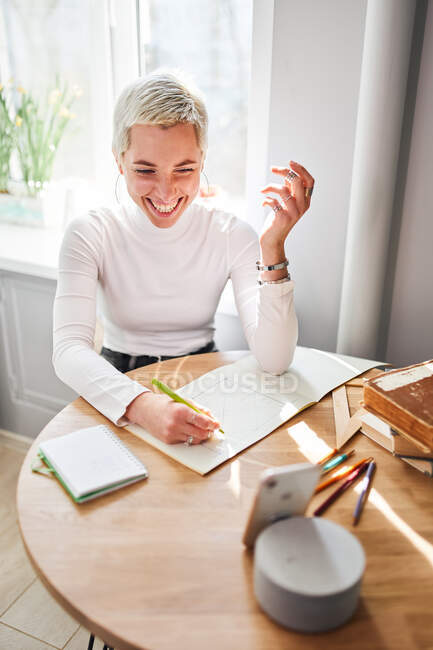Зверху щаслива жінка-астролог робить нотатки в блокноті за столом з чашкою кави вдома на сонячному світлі — стокове фото