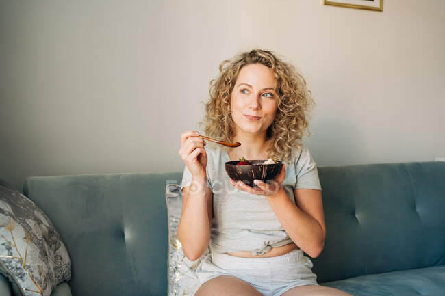 Веселая женщина в домашней одежде наслаждается вкусной едой в миске, сидя на удобном диване со скрещенными ногами с улыбкой — стоковое фото