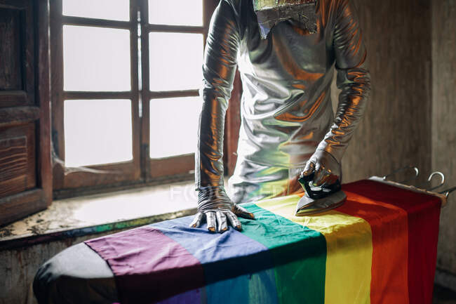 Persona irreconocible en traje de plata y caja en la cabeza planchando la bandera LGBTQ mientras está de pie en una habitación abandonada - foto de stock