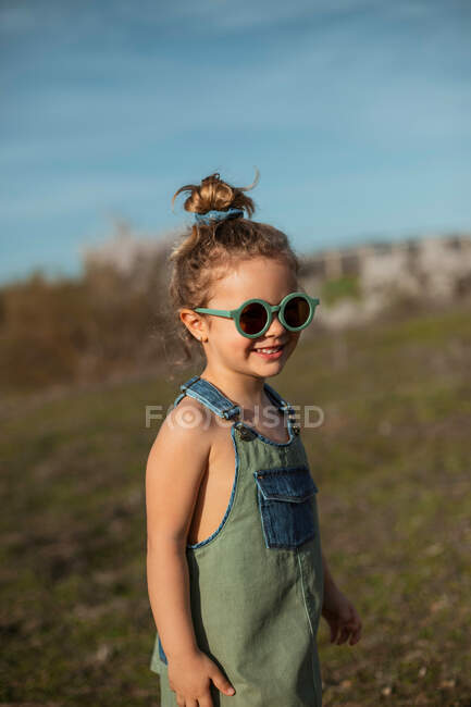 Conteúdo menina em macacão e óculos de sol em pé no prado e curtindo o verão no dia ensolarado no campo — Fotografia de Stock
