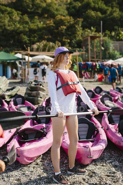 Mujer joven seria de cuerpo entero con chaleco salvavidas y gorra sosteniendo remo mientras está de pie en la costa cerca de muchos kayaks en un día soleado en Málaga España - foto de stock