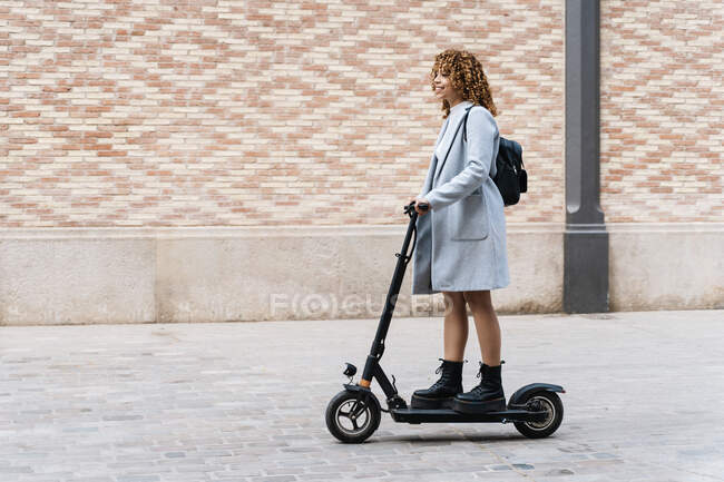 Full body content African American young female in casual wear riding scooter on paved sidewalk and looking away with smile — Stock Photo