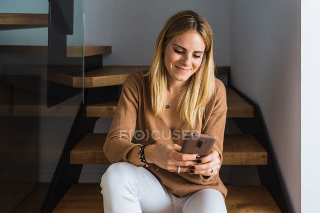 Счастливая женщина сидит дома на лестнице и смотрит на смартфон — стоковое фото