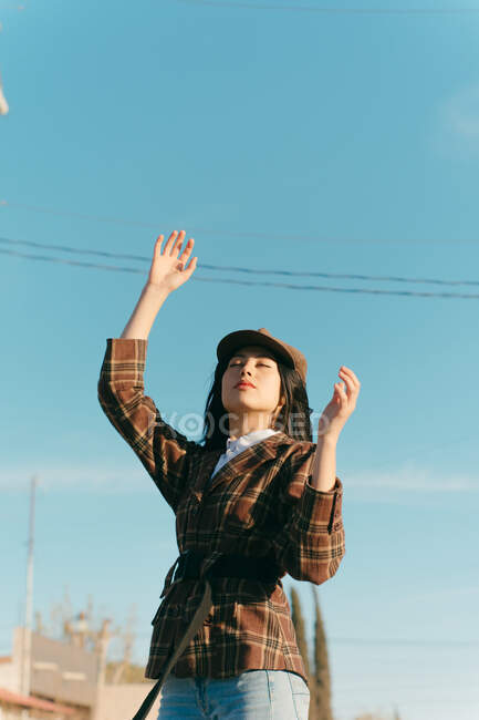 Young dreamy ethnic female in beret and checkered jacket standing under cloudy sky and looking up in sunlight — Fotografia de Stock