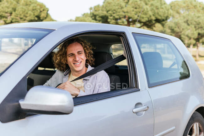 Happy young haired male looking away through open window of car while sitting at driver seat — Stock Photo