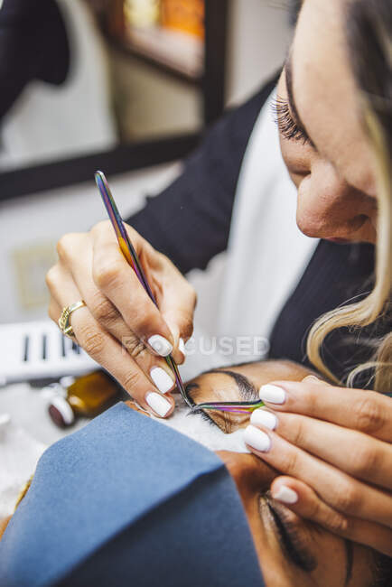 Crop unrecognizable cosmetologist with tweezers applying fake eyelashes for extension on eye of ethnic client with face protective mask in salon during coronavirus pandemic — Photo de stock