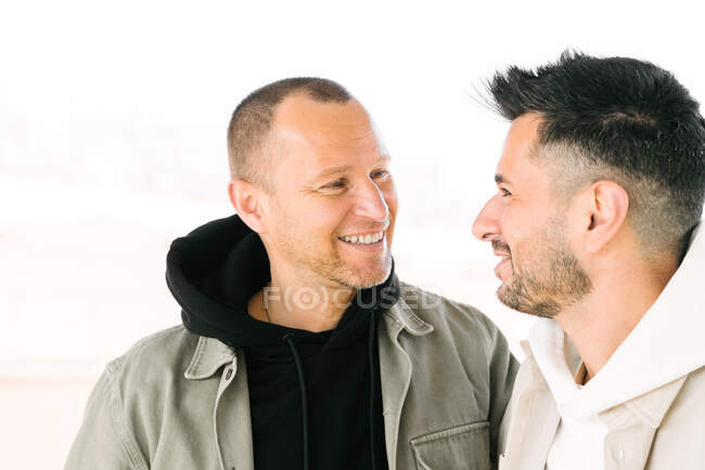 Side view of happy young multiracial gay couple hugging and looking at each other on street on sunny day - foto de stock