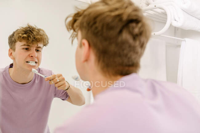 Vista posterior de un adolescente masculino concentrado con cabello de jengibre en camiseta cepillándose los dientes usando pasta de dientes y mirándose en el espejo en el baño - foto de stock
