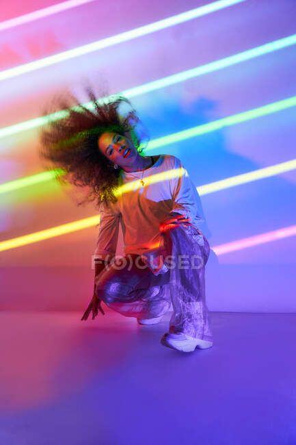 Corpo inteiro elegante confiante dançarina afro-americana com cabelo encaracolado e óculos de sol agachados olhando para a câmera em luzes de néon no estúdio de dança — Fotografia de Stock