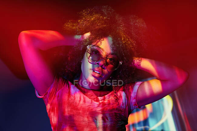 Jovem mulher afro-americana atraente em óculos de sol elegantes tocando o cabelo encaracolado e olhando para a câmera enquanto estava de pé em luzes de néon — Fotografia de Stock