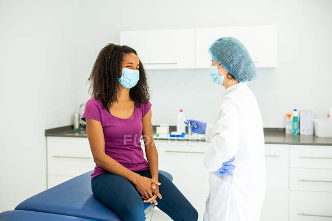 Cheerful female medical specialist in protective uniform, latex gloves and face mask talking to African American female patient before vaccinating in clinic during coronavirus outbreak — Stock Photo